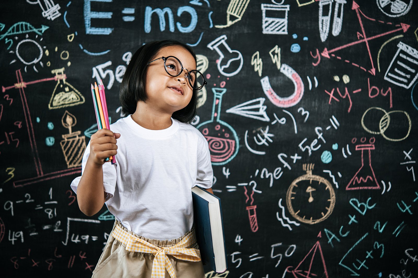 Selective focus, Little girl wearing eyeglasses holding colors pencil in hand and holding large textbook standing at front of blur blackboard in background, smile with happy